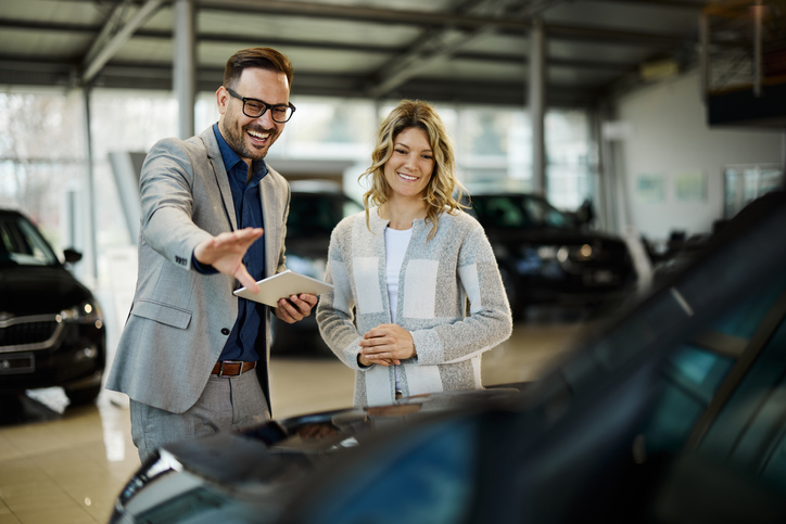 Happy Salesman Selling The Car To His Female Customer In A Showroom. - How Dealerships Can Sell More Cars And Get Mor...