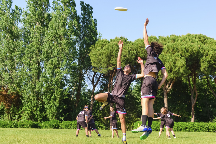 Group Of Young Teenagers People In Team Wear Playing A Frisbee Game In Park Oudoors. Jumping Man Catch A Frisbee To A...