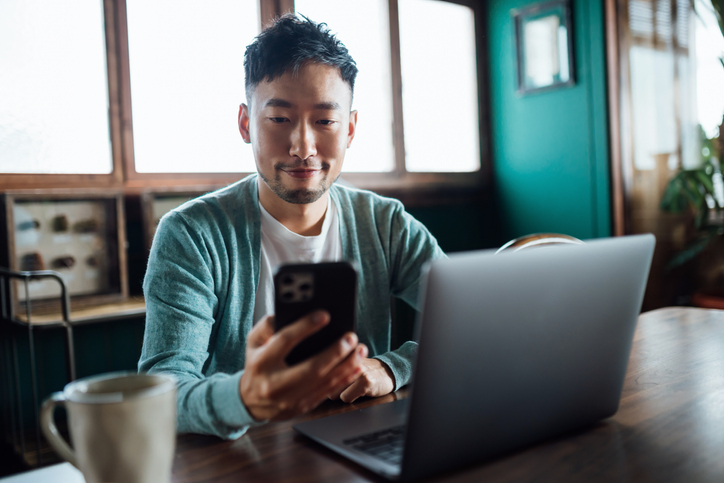 Confident Young Asian Man Looking At Smartphone While Working On Laptop Computer In Home Office - Is Social Media Pha...