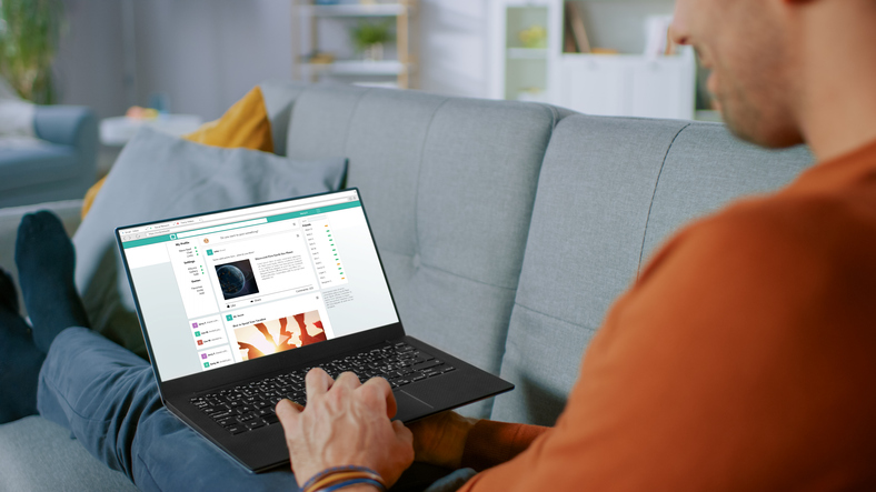 Handsome Young Man Sits At Home Uses Laptop And Rowses Through Social Network Feed Site. Over The Shoulder Shot Of An...