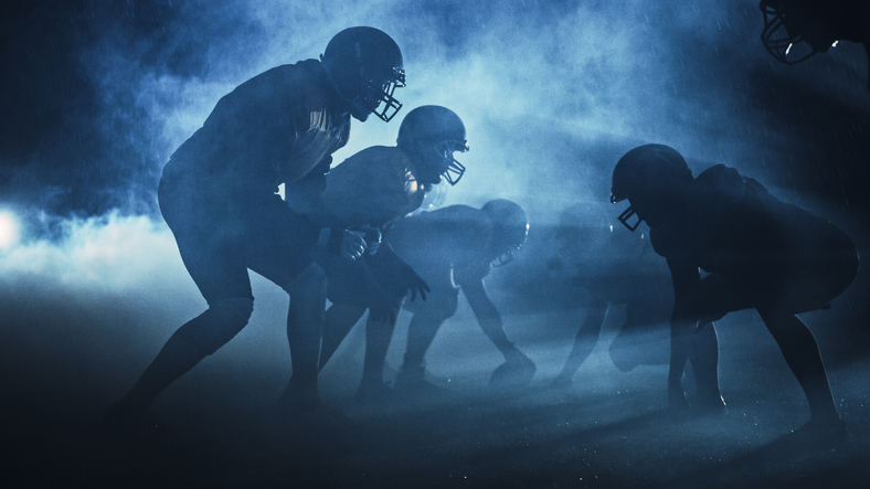 American Football Field Two Teams Compete: Players Pass, Run, Attack To Score Touchdown Points. Rainy Night With Athl...
