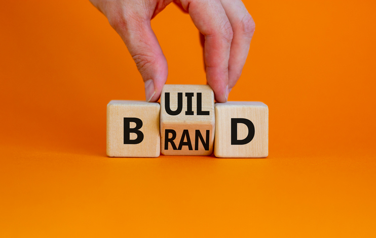 Build Your Brand Symbol. Businessman Turns Wooden Cubes And Changes The Word 'Build' To 'Brand'. Beautiful Orange Bac...
